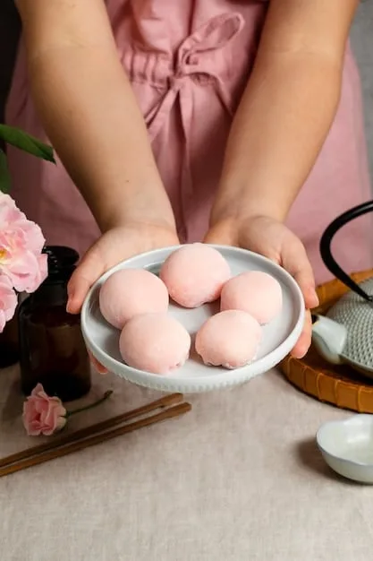 Close-up shot of a woman's hands carefully shaping mochi dough into small, round balls. The background shows a traditional Japanese kitchen with wooden bowls and a steamer.