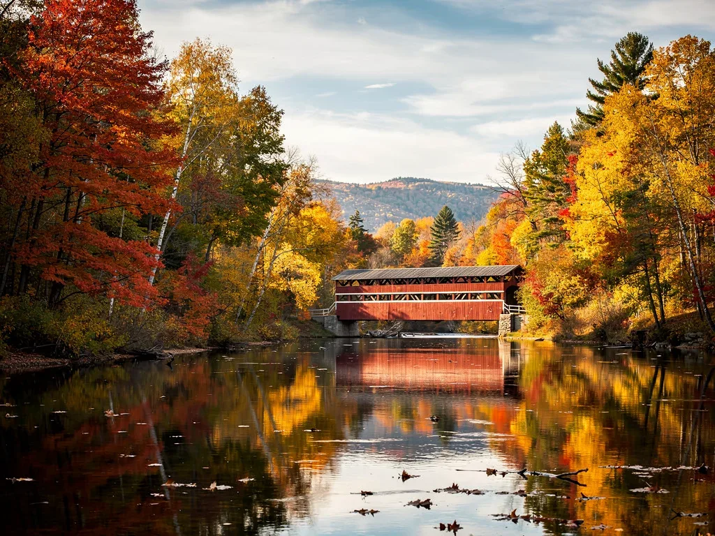 Historic covered bridge in Vermont framed by stunning peak autumn foliage colors and a serene river.