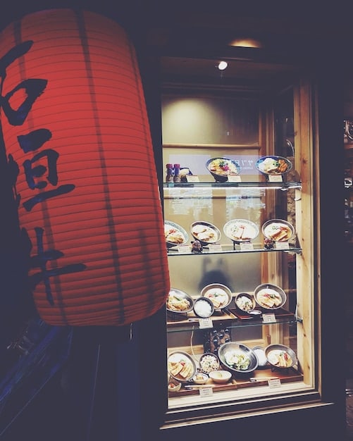 A close-up shot of a display window in a Japanese outlet mall. The window features a selection of high-end handbags and accessories from a luxury international brand. The items are neatly arranged, and the window display is well-lit, highlighting the quality and craftsmanship of the products. A small sign indicates a percentage discount.