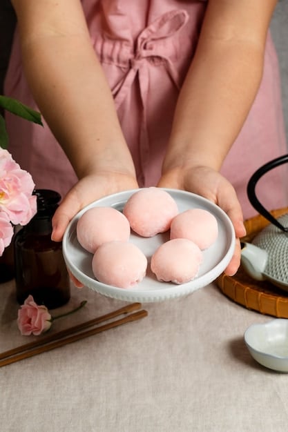 Close-up shot of a woman's hands carefully shaping mochi dough into small, round balls. The background shows a traditional Japanese kitchen with wooden bowls and a steamer.