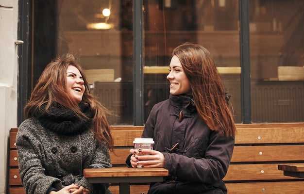 Two young women, possibly friends or sisters, are laughing and talking in a casual setting like a cafe or park. They convey a sense of closeness and warmth.
